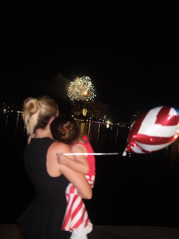 Mujer rubia de espaldas, peinado recogido, carga a niña de cabello castaño. Miran fuegos artificiales en cielo nocturno sobre agua y edificios. Globo rojo y blanco