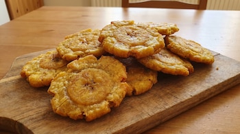 Primer plano de una docena de patacones colombianos fritos y aplastados, de color dorado, apilados sobre una tabla de cortar de madera clara.