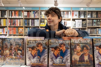 Bruno Pinasco, sonriendo con cabello rizado y suéter oscuro, se apoya en una mesa con su colección de libros 'Tea Shop' en una librería