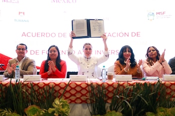 Cinco personas sentadas en una mesa, la mujer central de blanco levanta un documento abierto. Las otras cuatro personas aplauden. Al fondo hay un banner con texto y logos de Tijuana y San Felipe