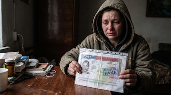 Una mujer con capucha llora mientras sostiene un documento que dice "VISA" y "REPUBLIKA FAKE" en una habitación oscura.