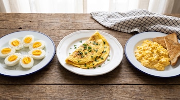 Platos con huevos duros cortados a la mitad, un omelet con perejil, huevos revueltos con pimienta y tostadas en una mesa de madera.