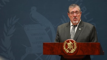 Un hombre con barba, anteojos y traje gris habla detrás de un podio de madera con el escudo de Guatemala
