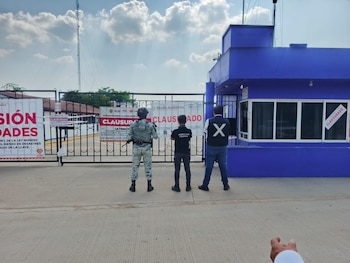 Tres hombres de espaldas frente a un portón metálico con carteles de clausura, un edificio azul con ventanas y un letrero de clausura, cielo nublado