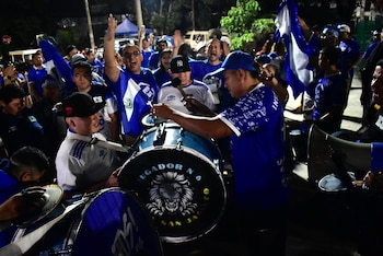 Varias personas con camisetas azules y blancas tocan tambores y ondean banderas de El Salvador durante la noche