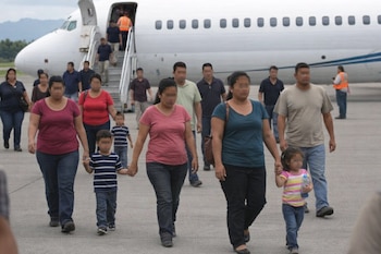 Un grupo de adultos y niños caminan sobre la pista de un aeropuerto. Al fondo, un avión blanco con pasajeros desembarcando por las escaleras.