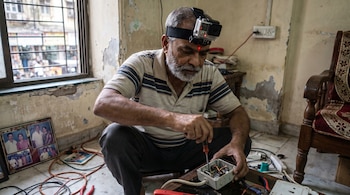 Hombre barbudo con cámara GoPro en la cabeza sujeta con banda, usa destornillador en una caja con cables en el piso, junto a ventana, marcos de fotos.