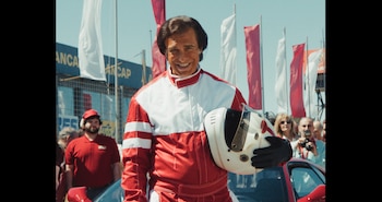 Un hombre con traje de piloto rojo y blanco sostiene un casco frente a banderas rojas y blancas en un autódromo