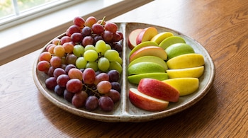 Un plato cerámico dividido contiene uvas rojas, verdes y moradas en un lado, y porciones de manzanas rojas, verdes y amarillas en el otro, sobre una mesa de madera.