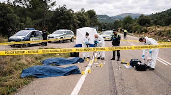 Dos cuerpos cubiertos con lonas azules yacen en la hierba al lado de una carretera acordonada con cinta amarilla de la policía y marcadores de evidencia.