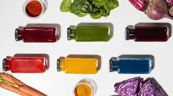 Vista aérea de seis botellas de zumos de colores (verde, rojo, azul, amarillo, morado) sobre blanco, con espinacas, remolachas, zanahorias, col morada y polvos.