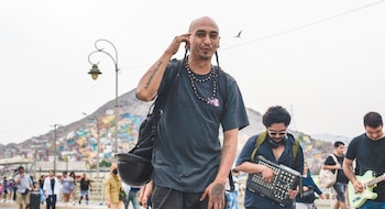 Un hombre calvo con barba y trenzas mira al frente mientras otros dos hombres tocan instrumentos, con personas y un cerro de casas de colores al fondo