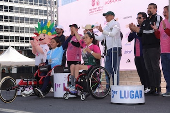 Dos mujeres en silla de ruedas con medallas y trofeos en un podio junto a hombres aplaudiendo y un disfraz de ajolote