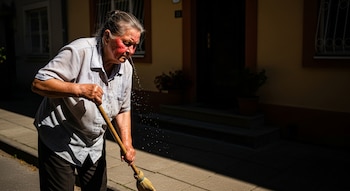 Se ve a una mujer mayor barriendo una vereda con una escoba. Su rostro está enrojecido y sudoroso, con gotas de agua cayendo. Viste una camisa clara.
