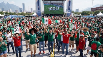 Una multitud de personas en un evento al aire libre celebra con banderas de México y carteles de gol frente a una pantalla gigante y edificios de una ciudad.