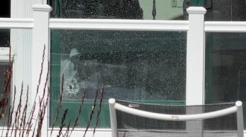 Un panel de vidrio agrietado con un orificio de impacto detrás de una barandilla blanca y el respaldo de una silla de exterior