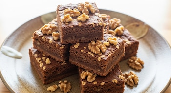 Una pila de brownies cuadrados de color marrón oscuro, espolvoreados con avena y nueces, servidos en un plato de cerámica beige con un fondo claro.