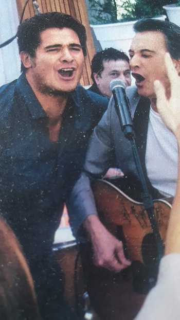 Dos hombres cantan frente a un micrófono, uno de ellos sosteniendo una guitarra acústica de madera