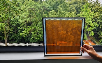 Una mano sostiene un panel rectangular semitransparente de color naranja con patrón de rejilla, frente a un ventanal con árboles verdes y una valla blanca