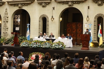 Hombre en podio, cinco personas en mesa, dos banderas de Guatemala, una bandera multicolor, arreglos florales, público, edificio de piedra con arcos