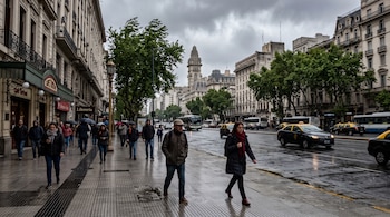 Transeúntes con abrigos caminan por la acera de una avenida con edificios tradicionales, árboles y vehículos en la calle mojada.