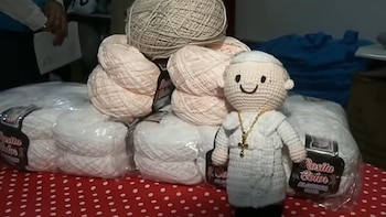Un muñeco tejido de color blanco junto a varios paquetes y ovillos de hilo sobre una mesa con mantel rojo de lunares blancos