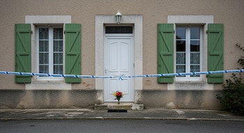 Fachada de casa rural con puerta blanca, dos ventanas con contraventanas verdes, cinta policial azul y blanca, y un ramo de flores en el umbral.