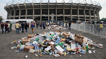 Montón de basura con botellas, cajas y envases en primer plano, con personas caminando y el Estadio Azteca de fondo bajo un cielo claro.