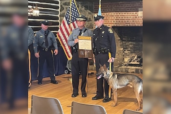 Oficiales de policía uniformados sostienen una placa, acompañados por un perro K9. Una bandera de Estados Unidos y una chimenea de ladrillos al fondo