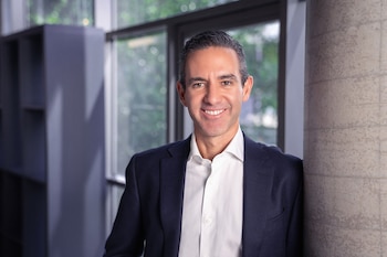 David Vélez viste un suéter morado y pantalón gris, de pie y con las manos en los bolsillos frente a ventanales de oficina