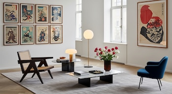 Salón con paredes blancas, ventanas grandes, un sillón de madera oscura y rejilla, dos mesas de centro, lámparas modernas, y obras de arte japonés enmarcadas en las paredes.