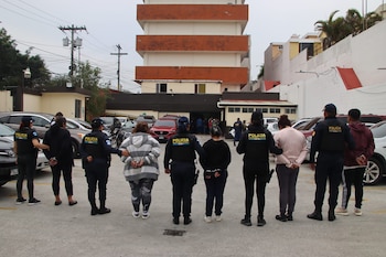 Oficiales de PNC Guatemala escoltan a personas detenidas, algunas con esposas, en un estacionamiento exterior con vehículos y edificios