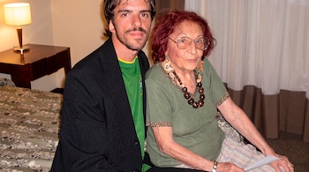 Un hombre joven con barba y saco junto a una mujer anciana con gafas y collar de madera sentados cerca de una cama