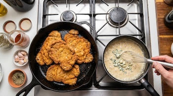 Vista cenital de milanesas doradas en una sartén de hierro fundido, junto a una mano batiendo salsa de roquefort en otra sartén, con especias y ajos cerca.
