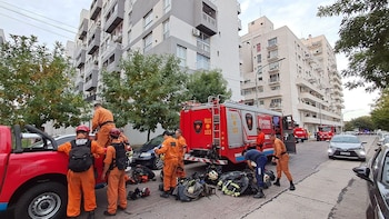 Vista de equipos de bomberos con uniformes naranjas y vehículos de rescate rojos en una calle con edificios residenciales altos, árboles y equipo en el suelo