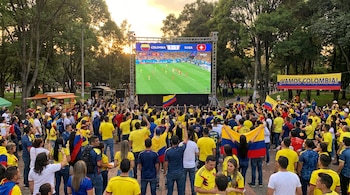 Multitud de personas con camisetas amarillas ven un partido de fútbol entre Colombia y Suiza en una pantalla gigante instalada al aire libre, con árboles y una bandera "Vamos Colombia!".
