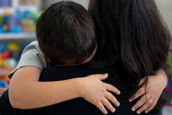 Vista posterior de un niño pequeño apoyando la cabeza sobre el hombro de una mujer con cabello oscuro a la que sostiene con un brazo.