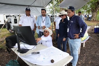 Cinco personas observan un monitor Dell sobre una mesa, un hombre con sombrero señala la pantalla, otro hombre sostiene un controlador de dron, un mapa