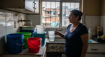 Karla Martínez en su cocina, recogiendo agua de un grifo que gotea en cubos de colores y jarras, con una ventana que muestra edificios urbanos.