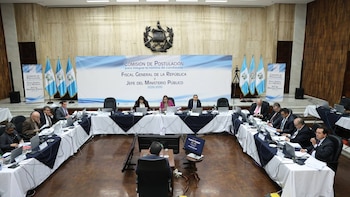 Sala de reuniones con mesa en U y varias personas sentadas con laptops y documentos. Al fondo, banderas de Guatemala y un letrero azul de "Comisión de Postulación" y el escudo
