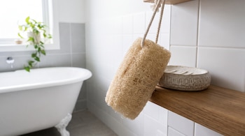 Una esponja vegetal cuelga de un estante de madera junto a una jabonera de cerámica, con una bañera blanca al fondo.