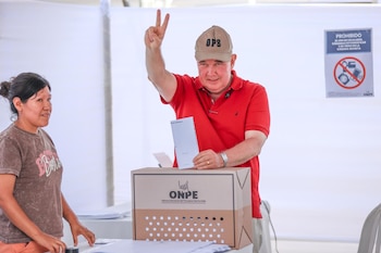 Hombre de polo rojo y gorra beige con la inscripción "OPS" sonríe mientras deposita un voto en una urna de la ONPE, haciendo el signo de la victoria con su mano derecha. Una mujer de camiseta marrón lo mira