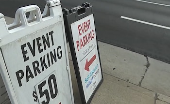 Dos letreros plegables de "EVENT PARKING" se encuentran en una acera de concreto junto a una calle. Uno muestra "$50" y el otro direcciona al SoFi Stadium