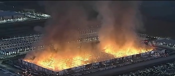 Vista aérea de un gran edificio industrial en llamas durante la noche, con densas columnas de humo que se elevan desde el techo