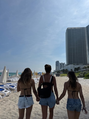 Tres mujeres vistas de espalda caminan tomadas de la mano por una playa de arena junto a tumbonas y grandes edificios