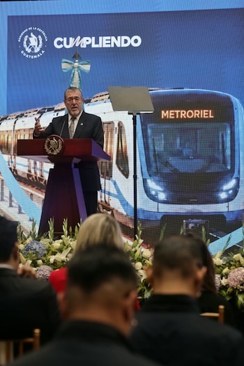 Bernardo Arévalo habla desde un podio frente a una pantalla que proyecta un tren con la palabra Metroriel, ante un público sentado