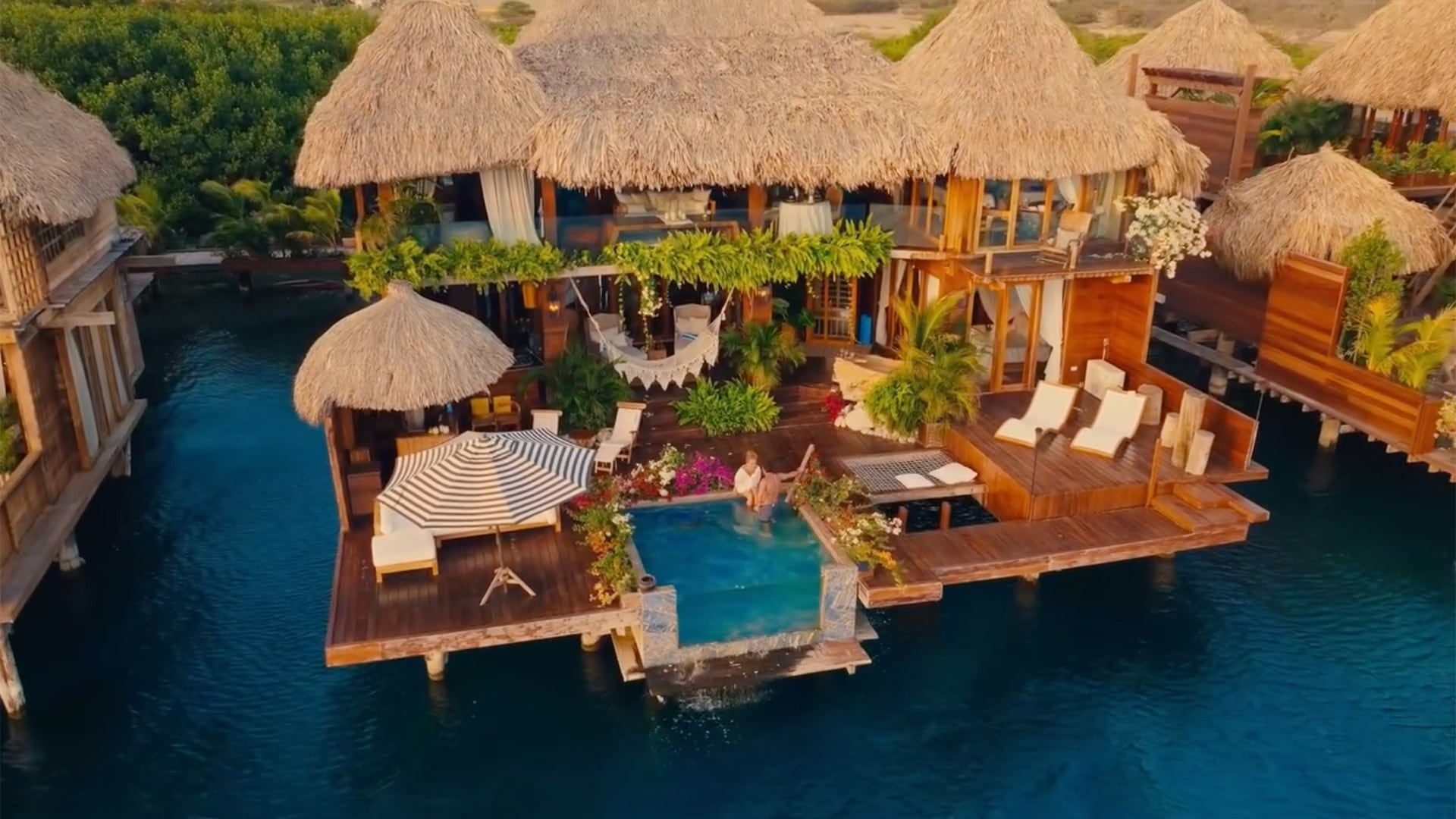 Vista aérea del exclusivo resort: la pareja se abraza en la terraza privada, enmarcada por techos de paja, hamacas y piscinas frente al mar Caribe