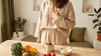 Una persona con ropa color beige se toca el pecho; sobre una mesa de madera hay brócoli, rodajas de naranja y kiwi, fresas, una taza de té con limón y un tazón con kéfir.