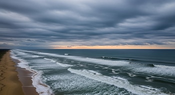 Vista aérea de una costa de arena con el océano presentando olas; el cielo está cubierto de nubes y el sol apenas se vislumbra en el horizonte.