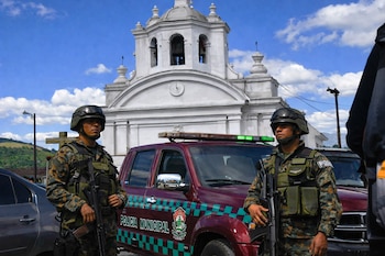 Dos agentes con uniforme camuflado, cascos y fusiles, junto a un vehículo de Policía Municipal rojo con luces. Al fondo, un edificio blanco con torres.